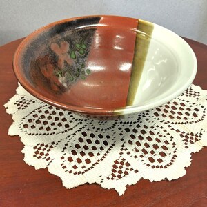 May include: A ceramic bowl featuring a multi-toned glaze with a deep red section, a dark brown area with green floral accents, and a smooth cream-colored side. The bowl has a wide, circular rim and a curved, deep basin design.