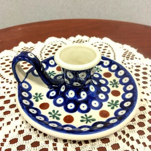 May include: A ceramic candle holder with a blue and white floral pattern. The holder has a circular base with a handle and a central cup for the candle. The design includes blue circles, green stars, and brown dots.