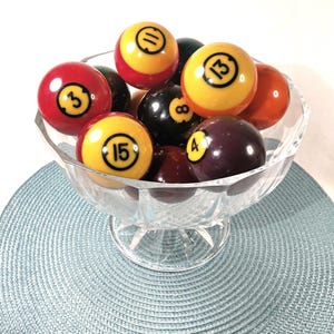 May include: A clear, faceted glass bowl filled with a collection of colorful billiard balls. The balls are numbered 3, 4, 8, 11, 13, and 15, featuring various solid and striped patterns in red, yellow, black, and dark purple hues.