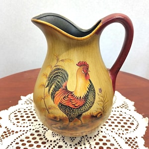 May include: A ceramic pitcher with a tan base and a dark red handle. The exterior features a painted rooster with a red comb, yellow neck feathers, and dark green and black body plumage, standing amidst stylized wheat and small floral elements.