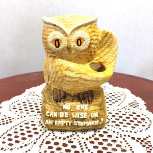 May include: A yellow ceramic owl figurine perched on a log base with brown accents and bright orange eyes. The owl holds a small circular opening in its wing. The log base features the embossed text: NO ONE CAN BE WISE ON AN EMPTY STOMACH.