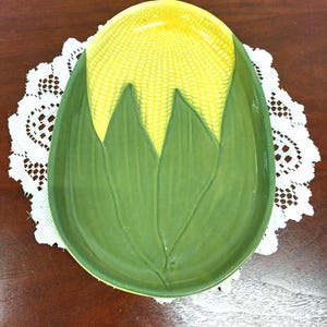 May include: An oval ceramic serving dish shaped like a corn cob. The top portion features a textured yellow pattern representing corn kernels, while the lower portion is molded into smooth, green, elongated leaves that extend toward the base.