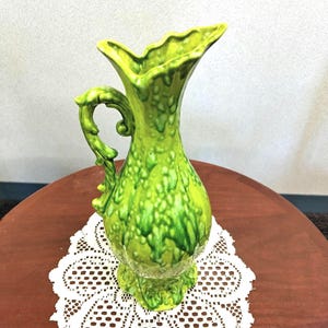 May include: A bright lime green ceramic pitcher featuring a textured, drip-glaze finish. The vessel has a flared, scalloped rim, a bulbous body, and an ornate, curved handle with intricate detailing. The base is wide and scalloped to match the rim.