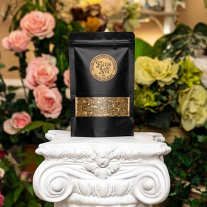 Pode incluir: Uma bolsa preta com um r&oacute;tulo dourado que diz "Elixir of Nyx Herbal Tea Blend" e cont&eacute;m ch&aacute; de folhas soltas. A bolsa est&aacute; sobre um pedestal branco.