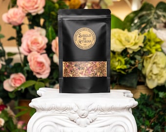 Może przedstawiać: Czarny woreczek z złotą etykietą z napisem "Shield of Athena Herbal Tea Blend" i mieszanką suszonych ziół i kwiatów.