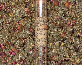 Może przedstawiać: Szklana probówka wypełniona suszonymi ziołami, z etykietą "Herbs of Olympus Super Herb Blood of Artemis Herbal Tea Net Wt 1 oz Relief & Balance".