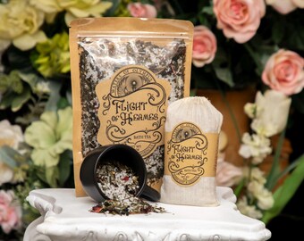 Może przedstawiać: Martwa natura z produktami do kąpieli. Czarny kubek wylewa herbatę do kąpieli na biały cokół. Przezroczysta torba i lniana torba z herbatą do kąpieli znajdują się za kubkiem. Torby mają etykiety z napisem "Waters of Olympus Flight of Hermes Bath Tea".