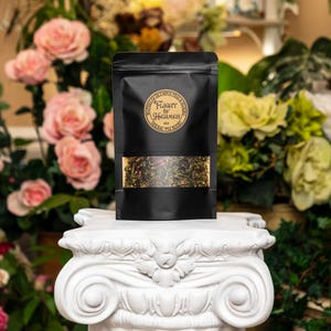 May include: A black pouch with a gold label that reads "Flight of Hermes Herbal Tea Blend". The pouch is filled with loose leaf tea and is sitting on a white pedestal.