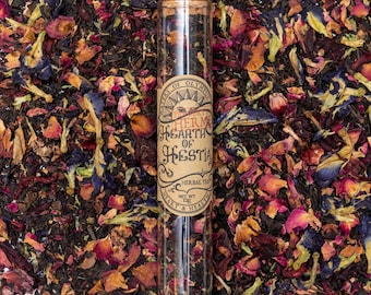Może przedstawiać: Szklana probówka wypełniona luźną herbatą, z korkowym korkiem. Probówka ma etykietę z napisem "Presents of Olympus, The Hearth of Hestia, Herbal Tea, Net Wt 1 oz, Heart & Health".