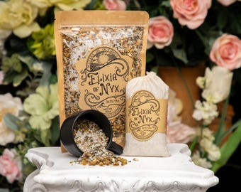 Może przedstawiać: Prezentacja produktów do kąpieli, w tym saszetka i mała torebka, z napisem "Elixia of Nyx". Czarny kubek jest przewrócony, wylewając herbatę do kąpieli na biały cokół. Tło przedstawia różowe i żółte kwiaty.
