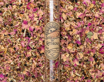Może przedstawiać: Szklana rurka wypełniona mieszanką suszonych ziół i kwiatów. Etykieta zawiera napis "Proper Herbs Shield of Athena Herbal Tea" z wzorem słońca i gwiazdy. Rurka leży na łóżku z suszonych płatków róż i innych ziół.