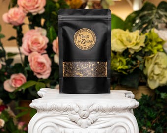 Może przedstawiać: Czarny woreczek z złotą etykietą z napisem "Tonic of Zeus Herbal Tea Blend". Woreczek jest wypełniony luźną herbatą i stoi na białym cokole.