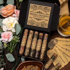May include: A black box set containing five glass vials of herbal tea blends. Each vial has a cork stopper and a label with text. The box lid reads "Sacred Herbal Blends Waters of Olympus Teas & Elixirs Crafted with Intent."