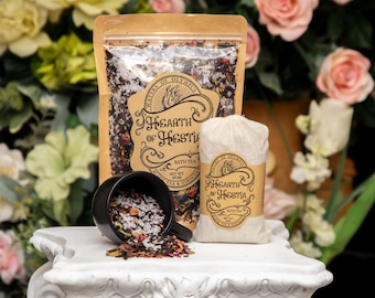 Może przedstawiać: Martwa natura przedstawia produkty do kąpieli. Papierowa torba kraft i mała płócienna torba są oznaczone jako "Hearth of Hestia". Czarny kubek wylewa herbatę do kąpieli na biały cokół. Tło wypełnione jest kwiatami.