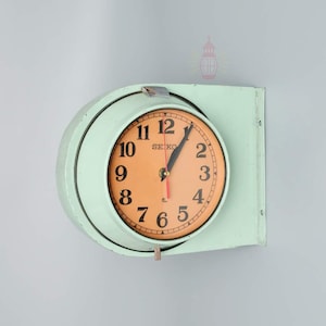 Op de afbeelding: Een vintage wandklok met een lichtgroen metalen frame en een oranje wijzerplaat. De wijzerplaat heeft zwarte cijfers en wijzers, met een rode secondewijzer. De klok is gemarkeerd met "SEIKO".