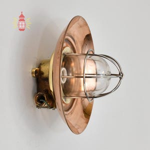 May include: A vintage wall-mounted light fixture with a copper shade and a brass base. The light has a clear glass globe encased in a metal cage. The fixture is designed for indoor or outdoor use.