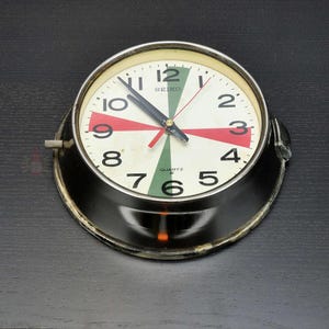 Könnte beinhalten: Eine Vintage Seiko Quartz Uhr mit weißem Zifferblatt, schwarzen Zeigern und einer schwarzen und silbernen Lünette. Das Zifferblatt der Uhr hat rote und grüne Akzente und schwarze Ziffern. Die Uhr befindet sich vor einem dunklen Hintergrund.
