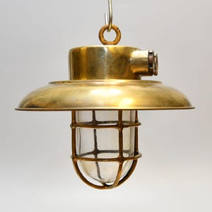 Op de afbeelding: Vintage messing hanglamp met een heldere glazen bol, omhuld door een metalen kooi. De lamp heeft een ronde kap en een ophanglus. Het messing heeft een warme, verouderde patina, wat de vintage oorsprong aangeeft.