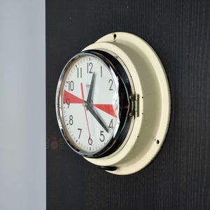May include: Cream-coloured Citizen clock with a black bezel and a chrome rim. The clock face is white with black numerals and hands, and red accents. The clock is mounted on a dark wall.