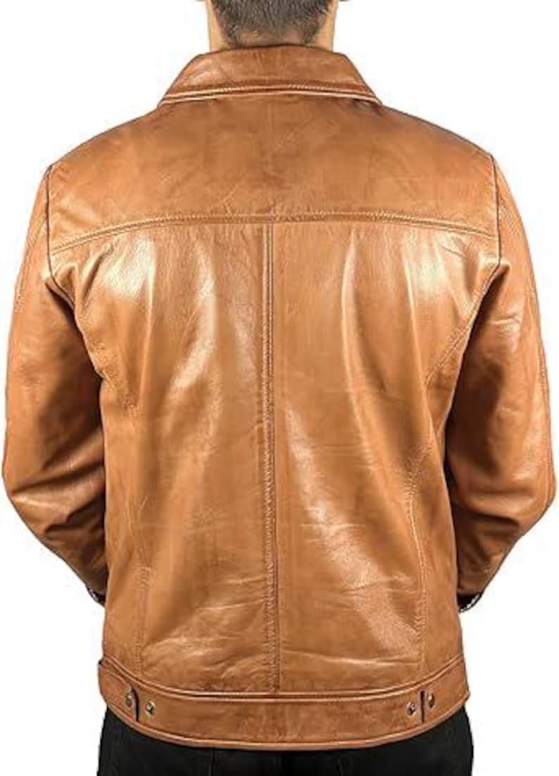 May include: A tan leather jacket with a classic design. The jacket features a collared neckline, a button-down front, and a tailored fit. The back of the jacket has a yoke detail and vertical seam. The jacket is made of a smooth, textured leather.