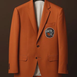 May include: A solid orange blazer featuring a notched lapel, two front buttons, and two flap pockets. A circular patch is sewn onto the left chest area, displaying the text BASNATRALL along with other indistinct lettering and a small graphic.