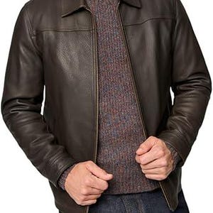 May include: A brown leather jacket with a collared neckline and a zip closure. The jacket is worn over a grey and brown speckled jumper. The person is wearing blue jeans.