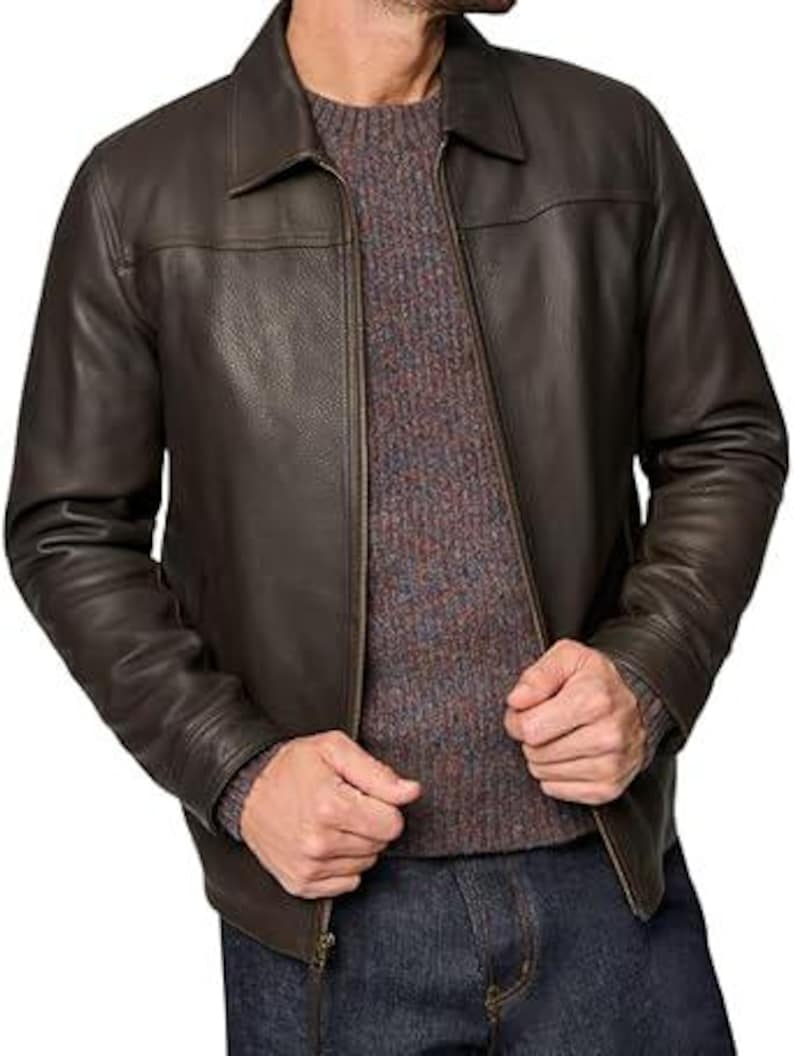 May include: A brown leather jacket with a collared neckline and a zip closure. The jacket is worn over a grey and brown speckled jumper. The person is wearing blue jeans.