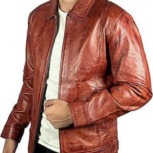 May include: A reddish-brown leather jacket with a collared neckline and a zip closure. The jacket has a classic design with a slightly distressed appearance. The model is wearing a white t-shirt underneath.