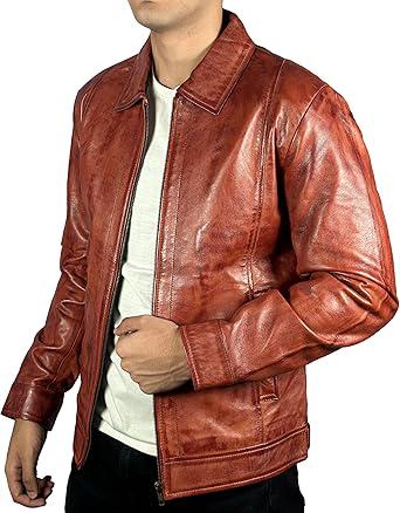 May include: A reddish-brown leather jacket with a collared neckline and a zip closure. The jacket has a classic design with a slightly distressed appearance. The model is wearing a white t-shirt underneath.