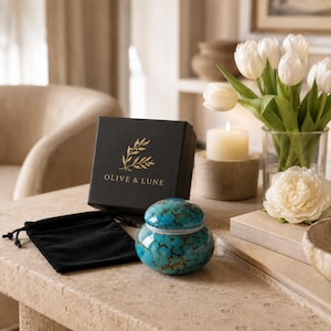 May include: A small, round, turquoise-colored jar with a lid featuring a dark brown marbled pattern, accompanied by a black drawstring pouch and a black square gift box with the gold-printed text OLIVE & LUNE and a leaf branch illustration.