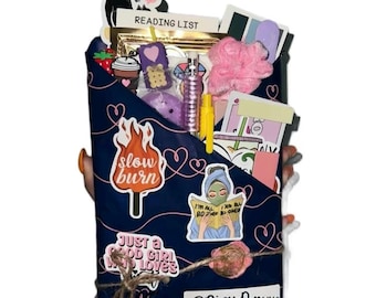 May include: A navy blue gift box with various items, including stickers with text like "slow burn" and "Spicy Romance." The box contains a pink loofah, a purple item, and a small bottle. The words "READING LIST" are visible.