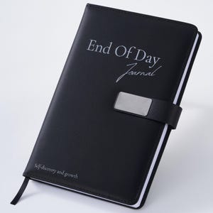 May include: Black journal with the text "End Of Day Journal" and "Self-discovery and growth" printed on the cover. The journal has a silver metal clasp and a black ribbon bookmark. The journal is closed.