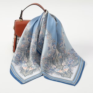 Op de afbeelding: Een lichtblauwe zijden sjaal met een bloemenpatroon, gedrapeerd over een bruine leren handtas. De sjaal heeft een gedetailleerd ontwerp met tinten blauw, roze en wit. De handtas heeft een handvat aan de bovenkant en een patroon aan de zijkant.