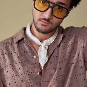May include: A person wearing black-framed sunglasses with yellow lenses and a white scarf. The person is wearing a button-down shirt with a geometric pattern in a muted mauve colour.