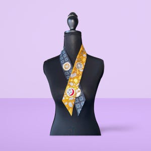 May include: A black and gold patterned silk scarf with a geometric design. The scarf is tied around a black mannequin's neck.