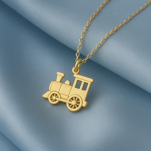 Puede incluir: Un collar con colgante de tren dorado. El colgante es una miniatura detallada de una locomotora clásica con ruedas, chimenea y ventanas. La cadena del collar es delicada y dorada, sobre un fondo azul.