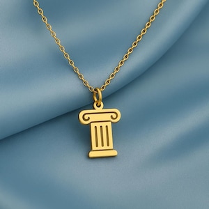 Puede incluir: Un collar dorado con un colgante en forma de columna clásica. El colgante tiene un diseño detallado con líneas verticales y una parte superior decorativa. La cadena es delicada, sobre un fondo de tela azul.