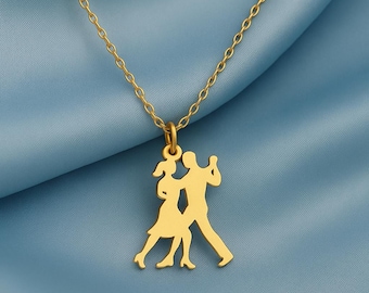 May include: A gold-colored necklace with a pendant depicting two dancing figures. The pendant is a silhouette of a man and woman in a tango pose. The chain is a delicate link style, and the background is a blue fabric.