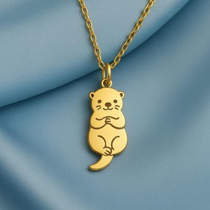 Pode incluir: Colar com pingente de lontra dourada. O pingente apresenta um design de lontra estilizado com detalhes gravados, suspenso por uma corrente delicada. A lontra é retratada com as patas juntas. O colar é exibido em um fundo azul.