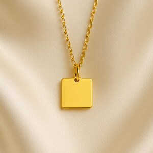 Pode incluir: Um colar dourado com um pingente quadrado. O pingente pende de uma corrente delicada, criando um design simples, mas elegante. O colar é exibido contra um fundo macio de cor creme.