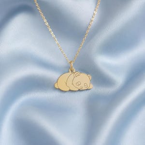 Op de afbeelding: Een goudkleurige ketting met een hanger in de vorm van een slapende pandabeer. De hanger hangt aan een fijne goudkleurige ketting. Het pandadesign bevat gegraveerde details voor de ogen, oren en gelaatstrekken op een glad oppervlak.