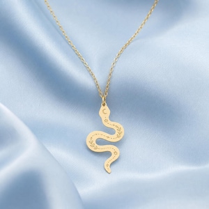 Può includere: Una collana con pendente a forma di serpente dorato. Il pendente presenta un design a serpente con un dettaglio di luna crescente e stella nella parte superiore. Il corpo del serpente è decorato con motivi a foglie e stelle.