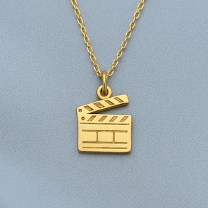 Peut inclure: Un collier doré avec un pendentif en forme de clap de cinéma. Le clap de cinéma présente des détails gravés et est suspendu à une chaîne délicate. Le fond est bleu clair uni.