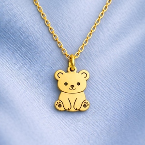 May include: A gold-coloured necklace with a teddy bear pendant. The bear is smiling and sitting. The chain is also gold, and the background is a light blue fabric. The pendant is a cute, cartoon-like design.