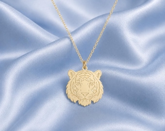 May include: A gold-colored necklace with a pendant shaped like a tiger's head. The pendant features detailed engraved lines depicting the tiger's facial features. The necklace hangs against a light blue satin background.