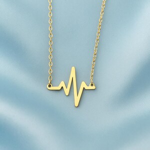 Op de afbeelding: Een goudkleurige ketting met een hanger in de vorm van een hartslag- of ECG-lijn. De hanger hangt aan een fijne ketting. De ketting wordt getoond tegen een lichtblauwe achtergrond.