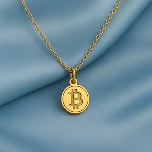 Op de afbeelding: Een goudkleurige ketting met een Bitcoin-symbool hanger. De hanger is een ronde munt met het Bitcoin "B" symbool in het midden. De ketting is delicaat en goudkleurig, tegen een blauwe stoffen achtergrond.