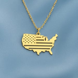 May include: A gold-colored necklace featuring a pendant shaped like the United States of America. The pendant has a cutout design depicting the American flag, with stars and stripes. The necklace hangs on a delicate chain against a blue background.