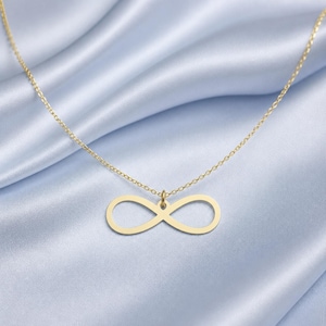 Puede incluir: Un collar dorado con un colgante con el símbolo del infinito. El colgante tiene forma de ocho estilizado. La delicada cadena cuelga sobre una tela de satén azul claro. Joyería.