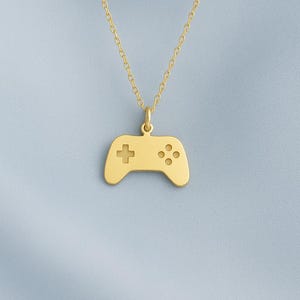 May include: A gold-coloured pendant necklace with a controller design. The pendant has a cross and four button details. The necklace chain is also gold-coloured, set against a light blue background.
